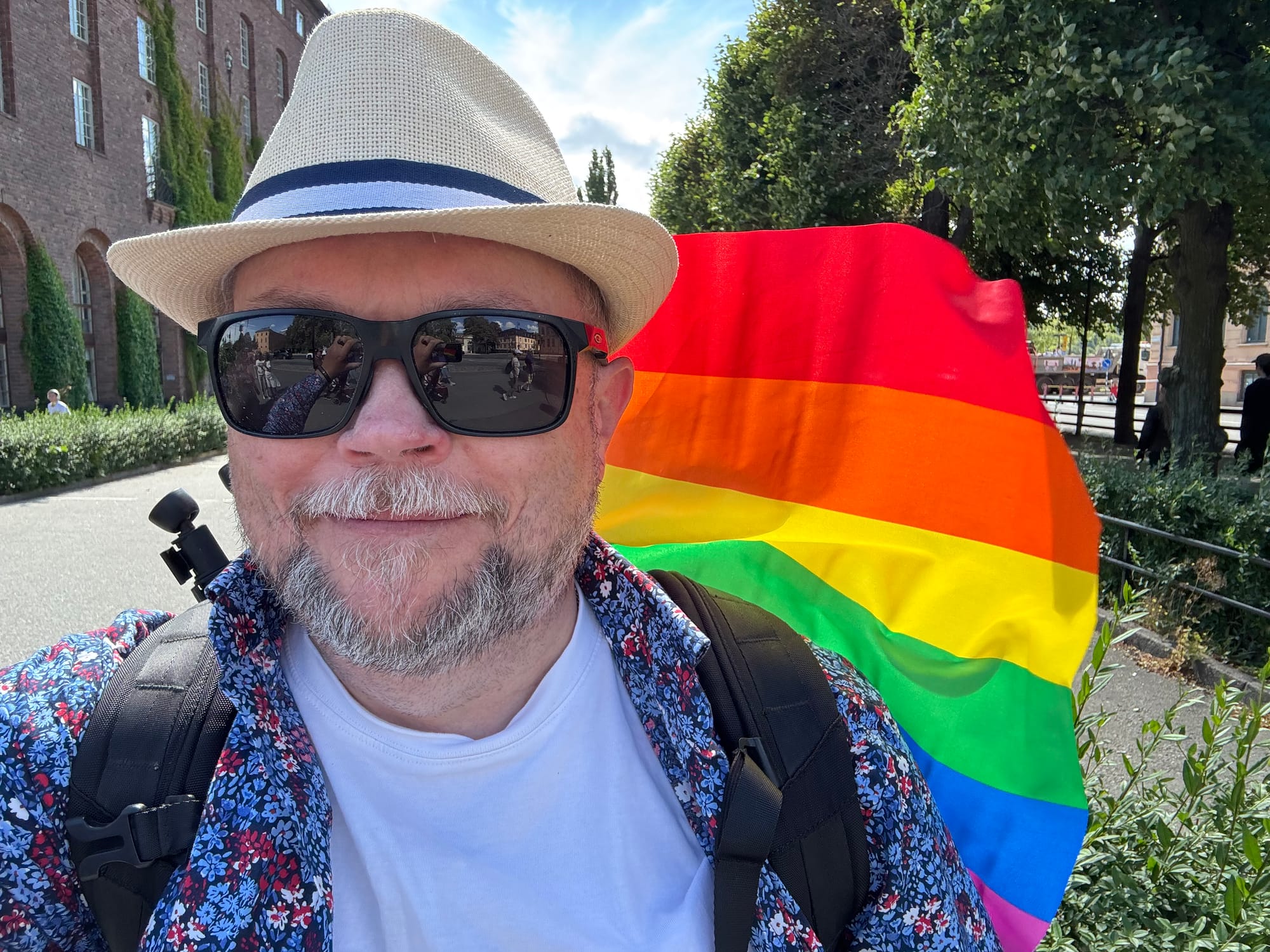 Per Axbom i hatt, solglasögon och blommig skjorta framför en vajande regnbågsflagga (som sitter i hans ryggsäck). Per står vid Stadshuset i Stockholm, som skymtar till vänster om honom.