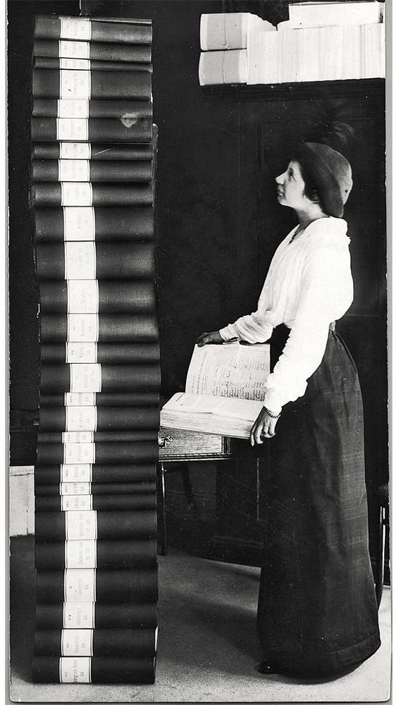 Black and white photo of a woman standing next to and looking at a stacked pile of about 30 binders of varying sizes. The stacked pile is around half a meter taller than her. She is holding a single binder open, giving an indication of how the binders were filled with signatures across all pages.