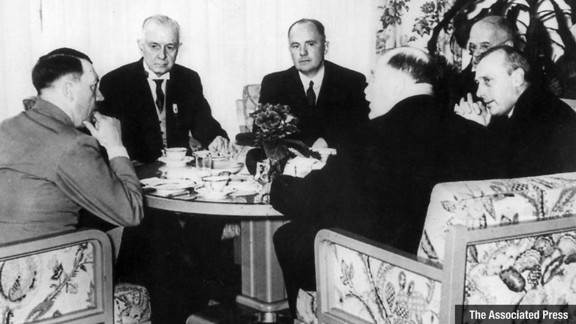 Black and white photo with six men seated in armchairs around a round coffee table - all gazes directed towards Hitler who is seated to the left in the picture. The people in the photo are, from left to right: Adolf Hitler; Thomas J. Watson; R. Schmidt; A. Frohwein; Van Vlissingen; Sir Arthur Balfour. (the order of the last four people is a bit unclear). Photo via Alamy/Associated Press. The picture is marked as taken July 12, 1937
