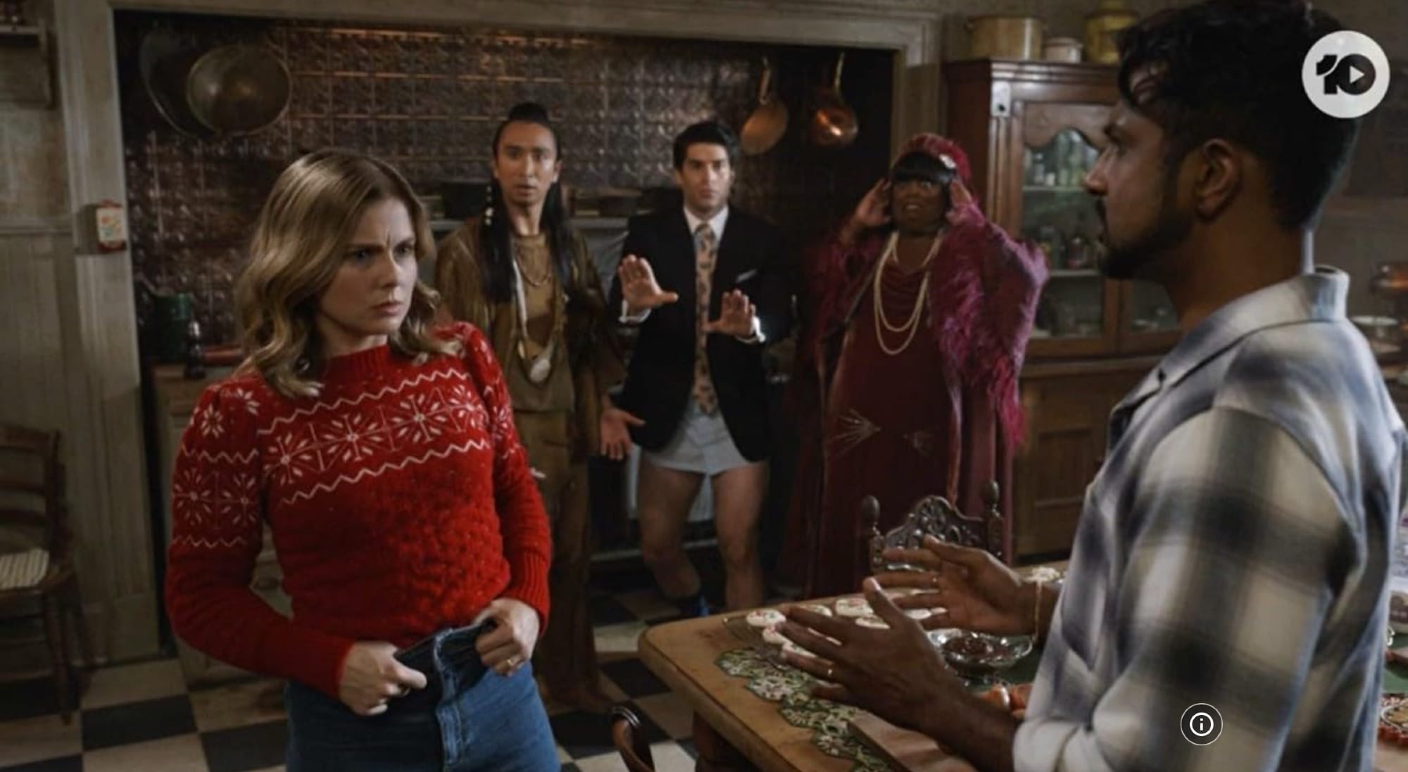 Samantha in American tv-series Ghosts, played by Rose McIver, in a red Christmas sweater talking to her husband Jay in the kitchen. Samantha has her thumbs hooked into her jeans, indicating that this is a scene where she is actually possessed by Thorfinn, the viking ghost. In the background, looking distressed, are the three ghosts Sass, Trevor and Alberta.