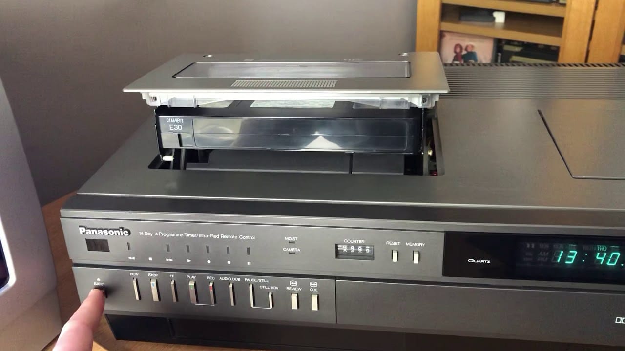 A top-loaded video cassette player with the cassette being ejected - also showing a finger pressing the eject button.