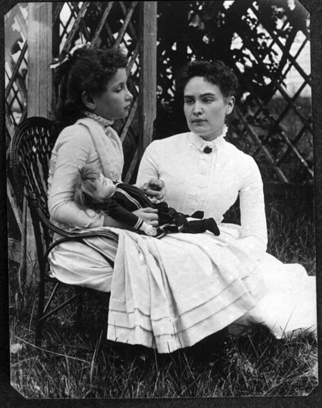 Helen Keller and Anne Sullivan in 1888, sitting outside. Helen is holding a doll, sitting in a low chair. Anne is sitting on the grass beside Helen, looking at her. Both are in long dresses.