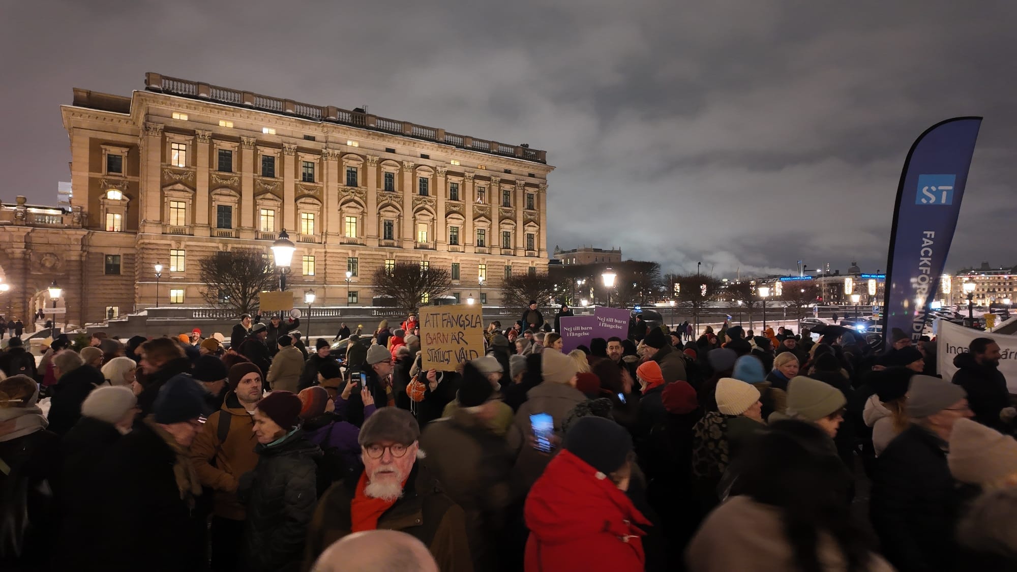 Folkmassa på Mynttorget i vinterkyla med riksdagshuset i bakgrunden. Handskriven protestskylt syns i mitten: Att fängsla barn är statligt våld. Och två mindre till höger: Nej till barn i fängelse.