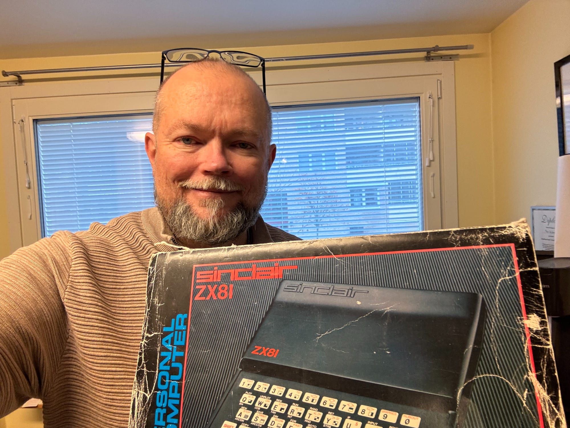 Per Axbom (middle-aged white guy with glasses pushed to the top of his bald head, sporting a greying beard) smiling and holding in front of him an old, worn box with a picture the Sinclair ZX81 computer.